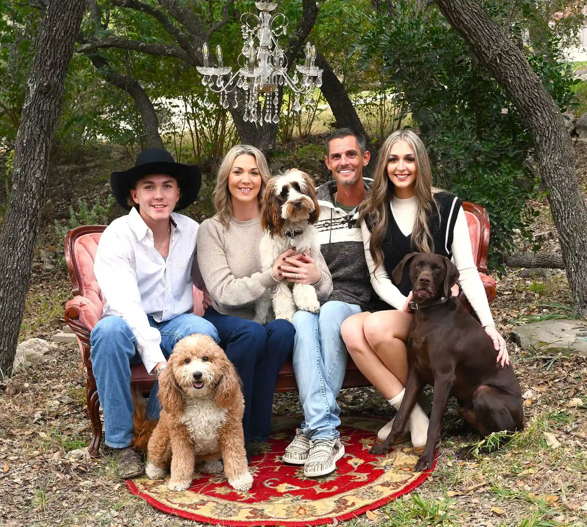 A family of five posing on a vintage couch outdoors, with two dogs sitting in front. The family consists of a young man, a woman, a man, and two teenagers, one holding a dog.

