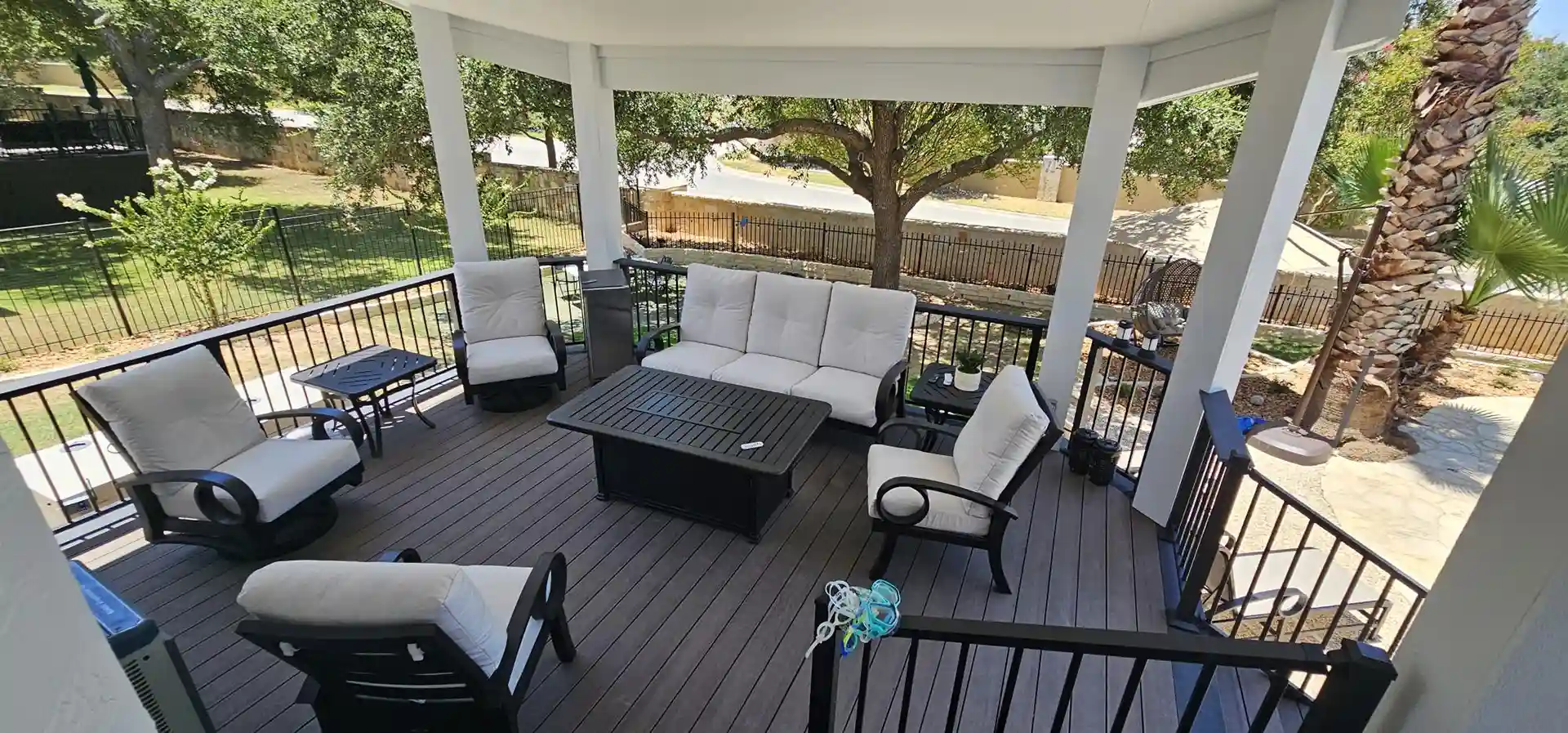 Covered outdoor deck with cushioned seating, table, and black railings overlooking a backyard with trees and a fence.
