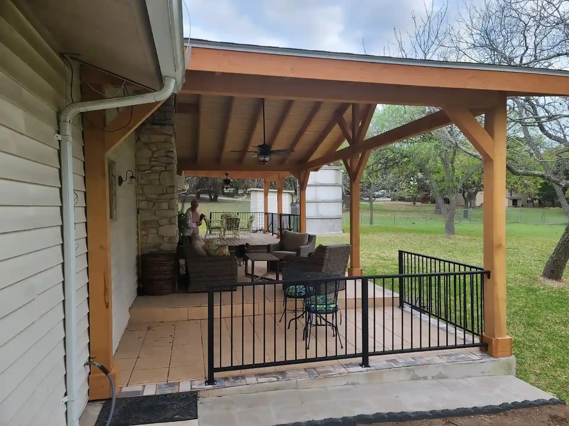 Covered outdoor patio with wooden beams, ceiling fans, and cushioned seating overlooking a grassy backyard.
