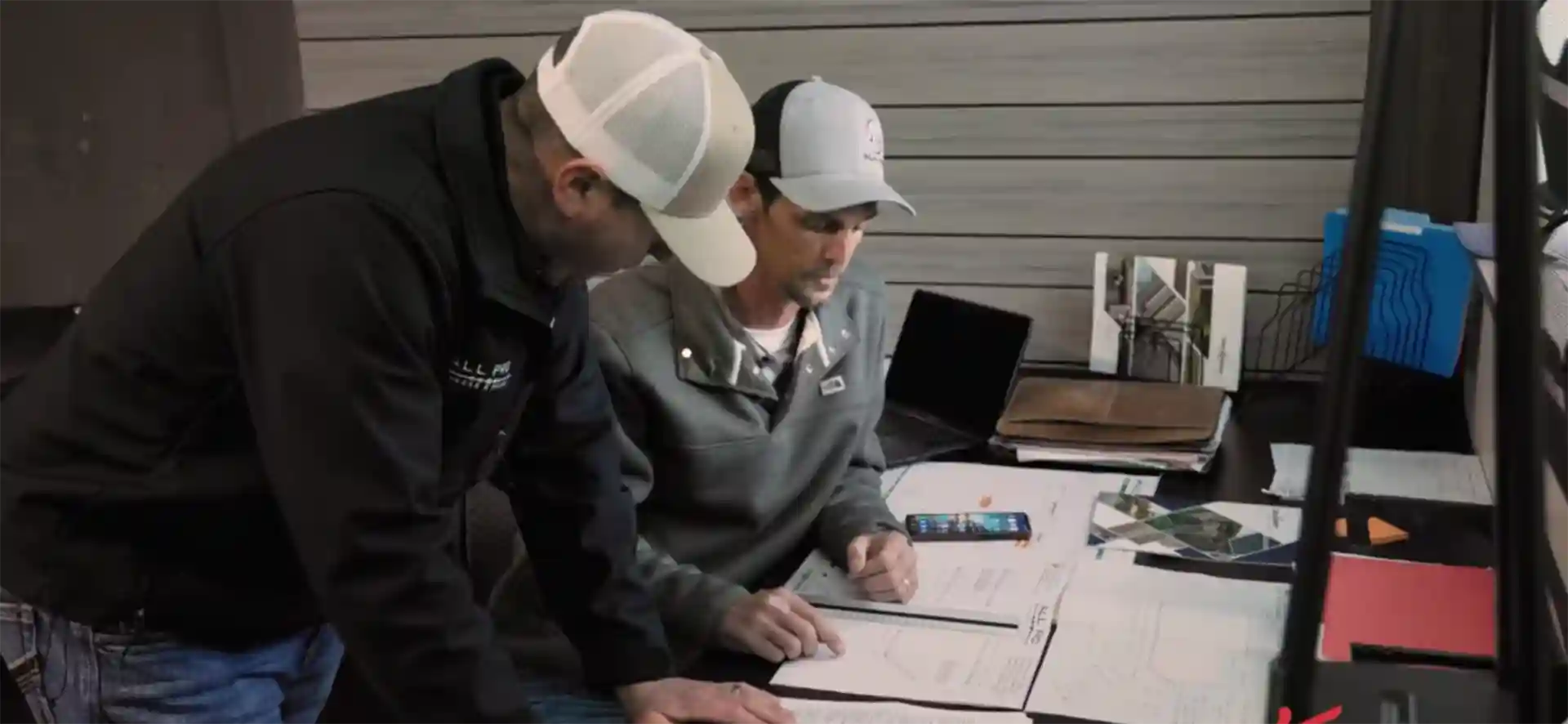 Two professionals reviewing deck design plans at a workspace with blueprints and a laptop.
