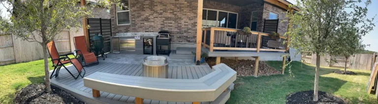 Outdoor deck area with built-in bench seating around a metal firepit, two orange lounge chairs, and an outdoor kitchen with grill and refrigerator, adjacent to a brick house and covered porch.