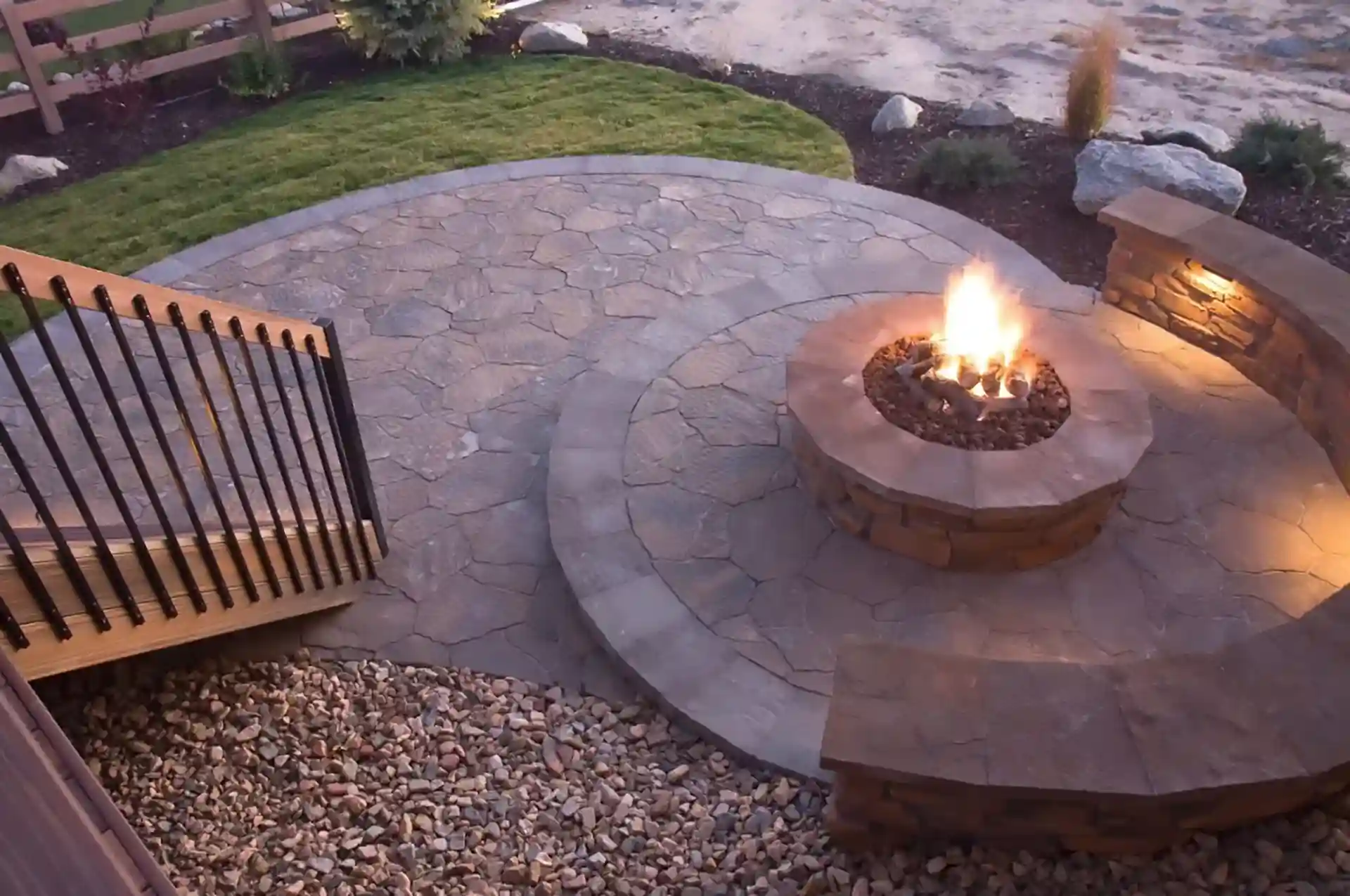 Stone patio with circular built-in firepit surrounded by curved stone seating walls, illuminated at dusk with landscape lighting and adjacent to wooden stairs with black metal railing. 