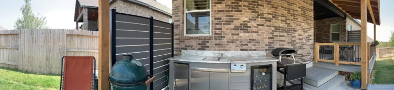 Modern outdoor kitchen featuring stainless steel appliances, a green ceramic grill, a wine cooler, and a pizza oven next to a brick wall.