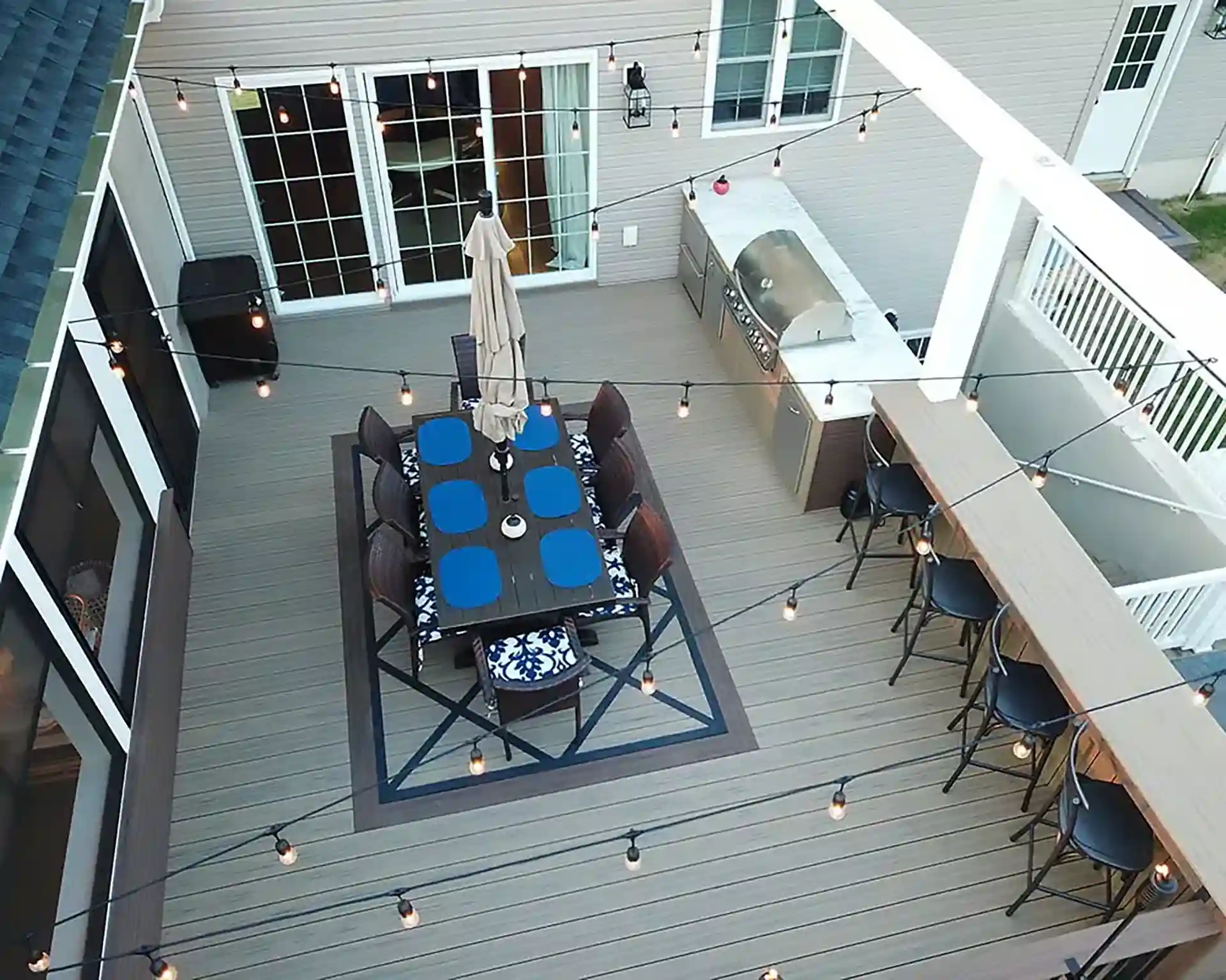 Overhead view of a modern outdoor deck with a dining table, built-in grill, bar seating, and string lights.
