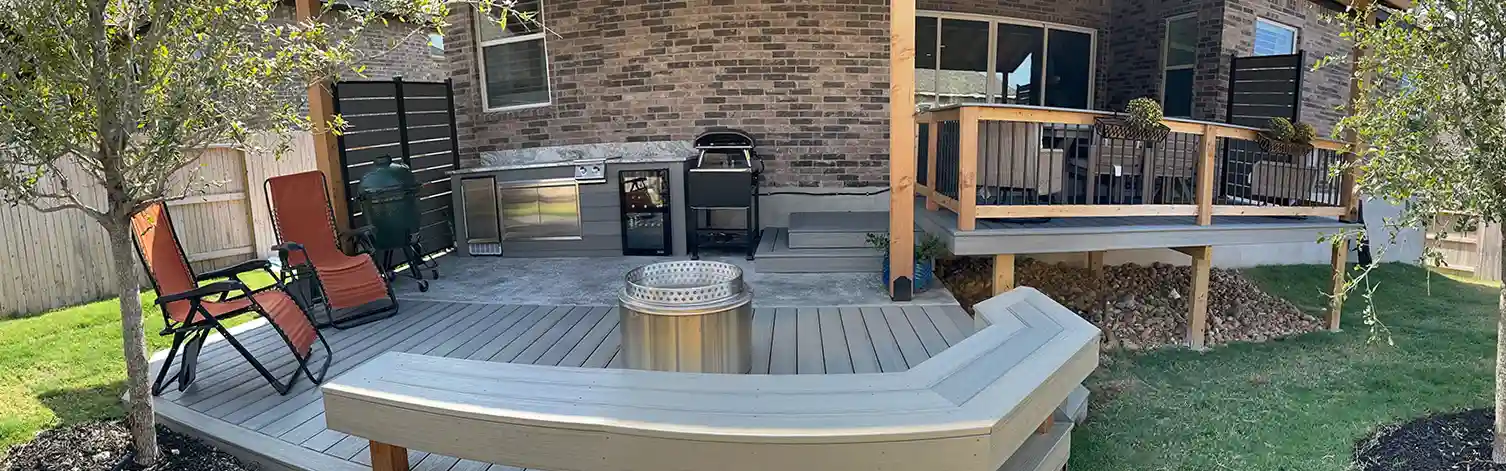 Outdoor kitchen and seating area on a raised deck with built-in grill, fire pit, and covered patio attached to a brick house.

