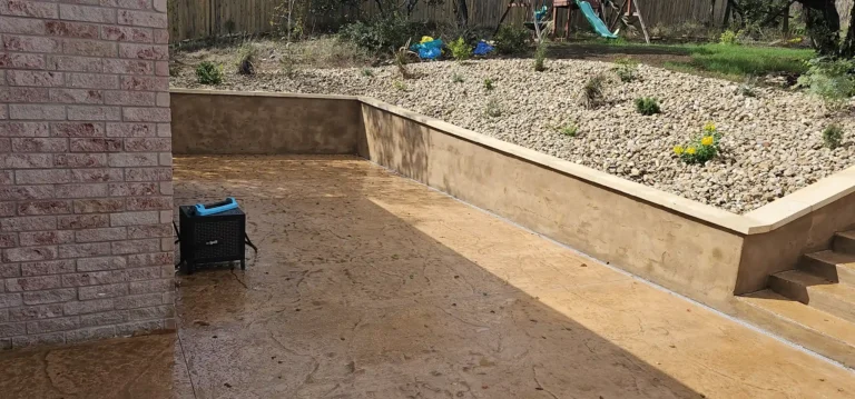 A brown, stamped concrete walkway is pictured. The walkway extends from a brick wall on the left into an area with a retaining wall and a rocky, sloped surface on the right.