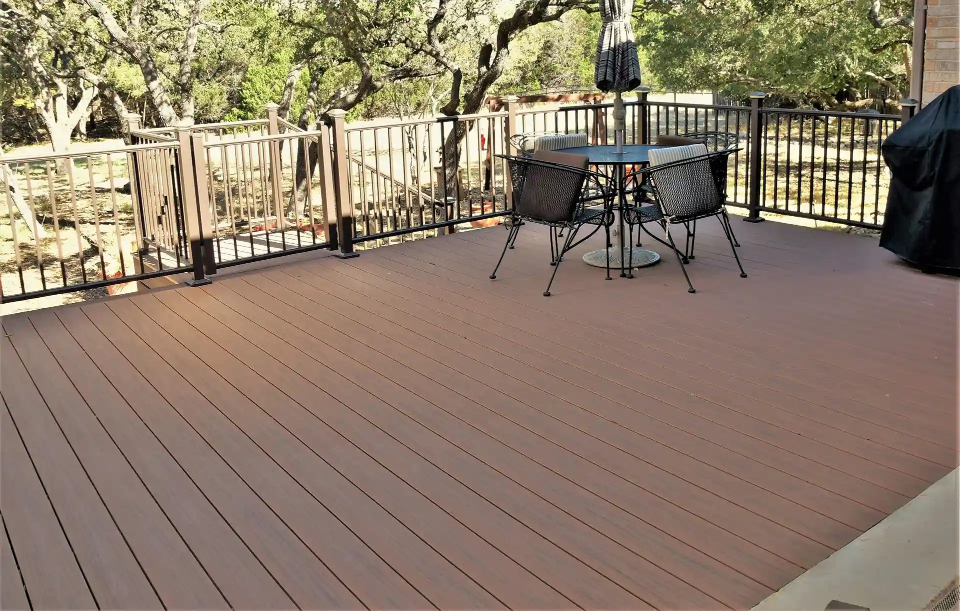 Spacious composite deck with brown decking boards, black metal railing, and outdoor dining set, surrounded by trees and natural landscape.
