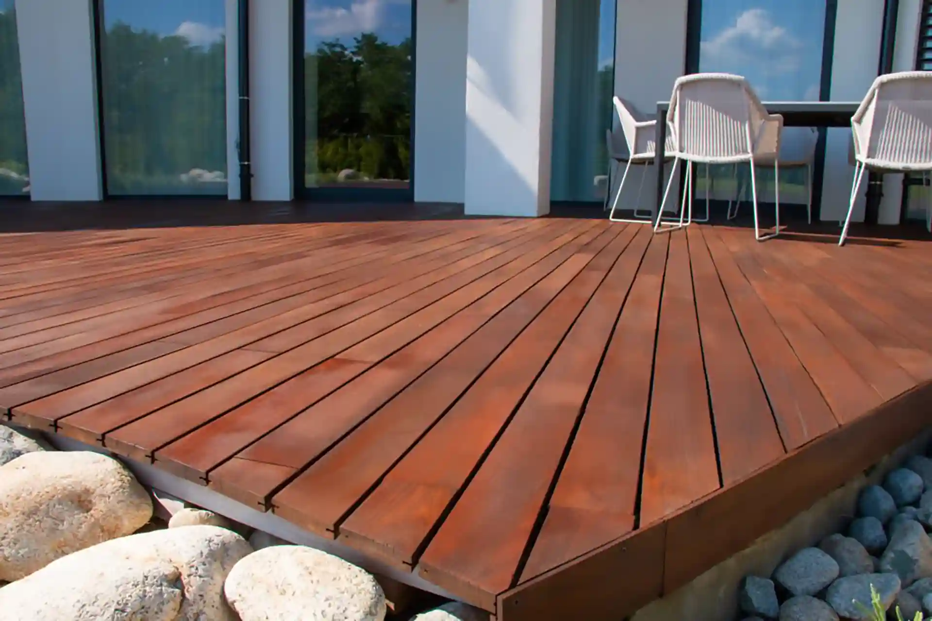 Modern outdoor deck built with rich brown IPE hardwood boards, featuring clean lines and a smooth finish, with patio furniture placed near glass doors.
