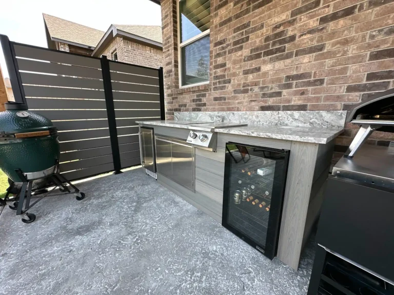 A modern outdoor kitchen features a stainless steel grill, wine cooler, and gray stone countertop set on a textured stamped concrete surface.