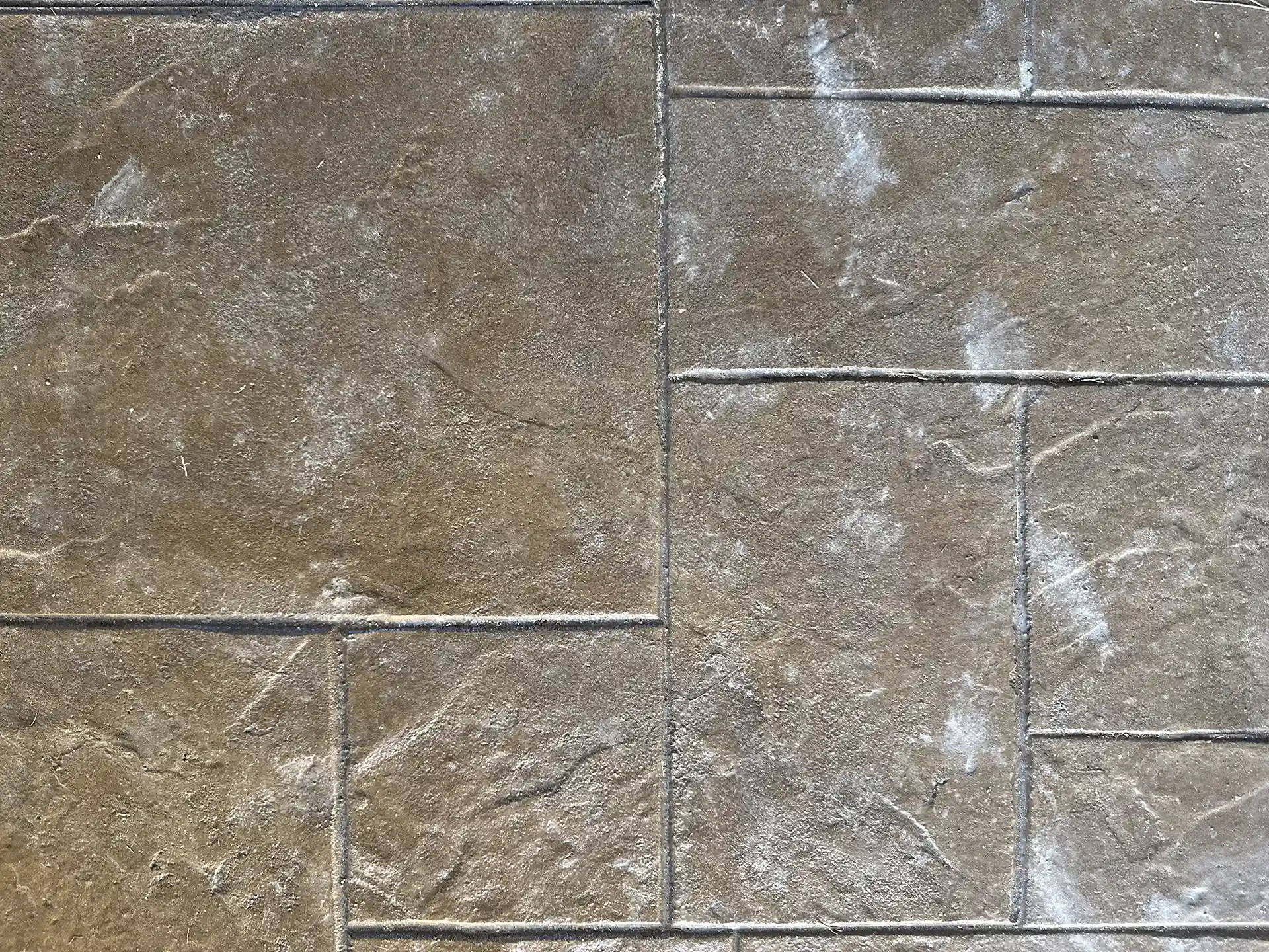 Close-up of a stamped concrete surface with a textured pattern resembling stone tiles. The surface has a rich, brown color with subtle variations.
