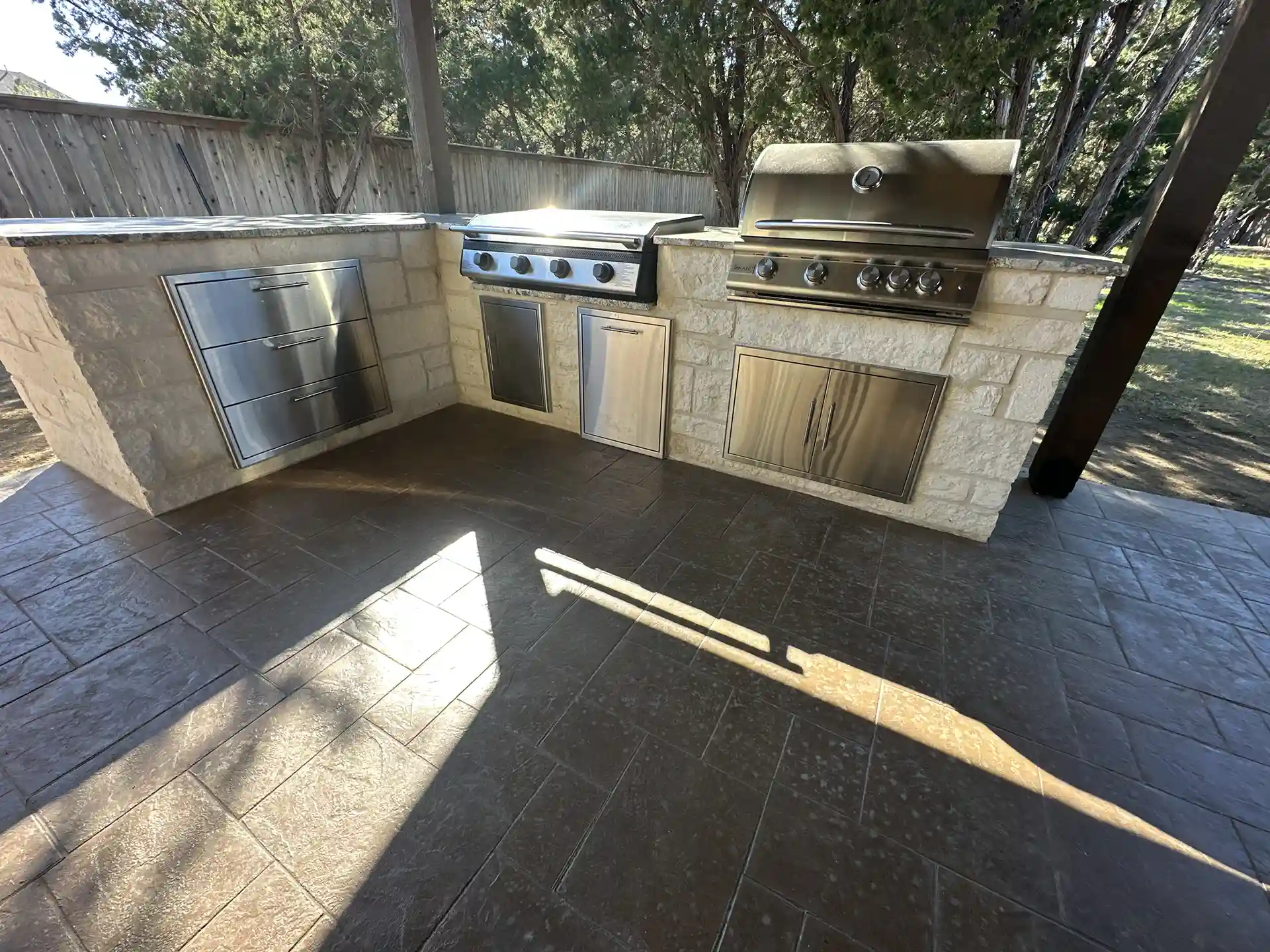 An outdoor kitchen featuring steel appliances, including a grill, drawers, and storage, is set on a stamped concrete floor with a textured, stone-like pattern.
